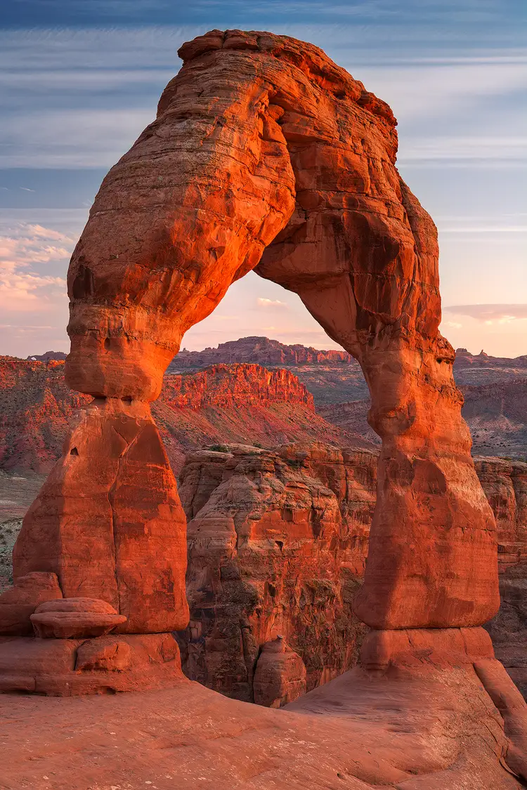 Photography location; In this vertical image Delicate Arch in Arches National Park fills the frame and is softly side lit by orange light from the setting sun. Framed inside the egg shaped opening of this tall arch one see three layers of rocky ridges cresting in the middle and also glowing orange from soft, sunset side light. Only a small amount of orange, clouded sky is between the top ridge and the bottom of the arch. The remaining sky, on the outside of the arch, is filled with horizontally striated clouds that are white high up and light orange near the horizon.