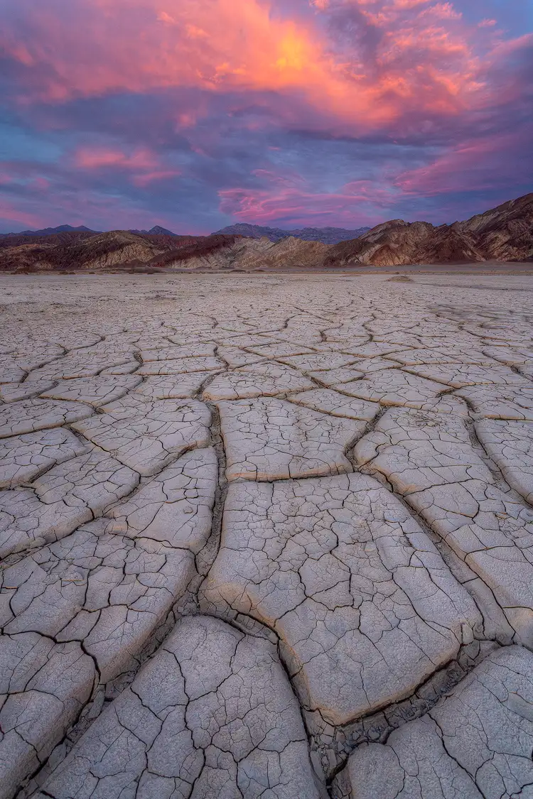 Death Valley workshop location. A vertical image of small mud cracks sitting atop other, much larger, mud cracks. The web like lines between the larger mud cracks create powerful visual elements that sweep the viewer from the foreground to the desert mountain background, and above to the swirling pink and yellow sunset clouds set against a blue sky.