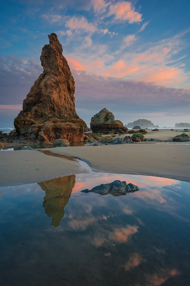 Photography location; Vertical, sunrise photo of pink clouds and a blue sky surrounding a pointy rock and reflecting in a tide pool on Bandon Beach, Oregon coast.