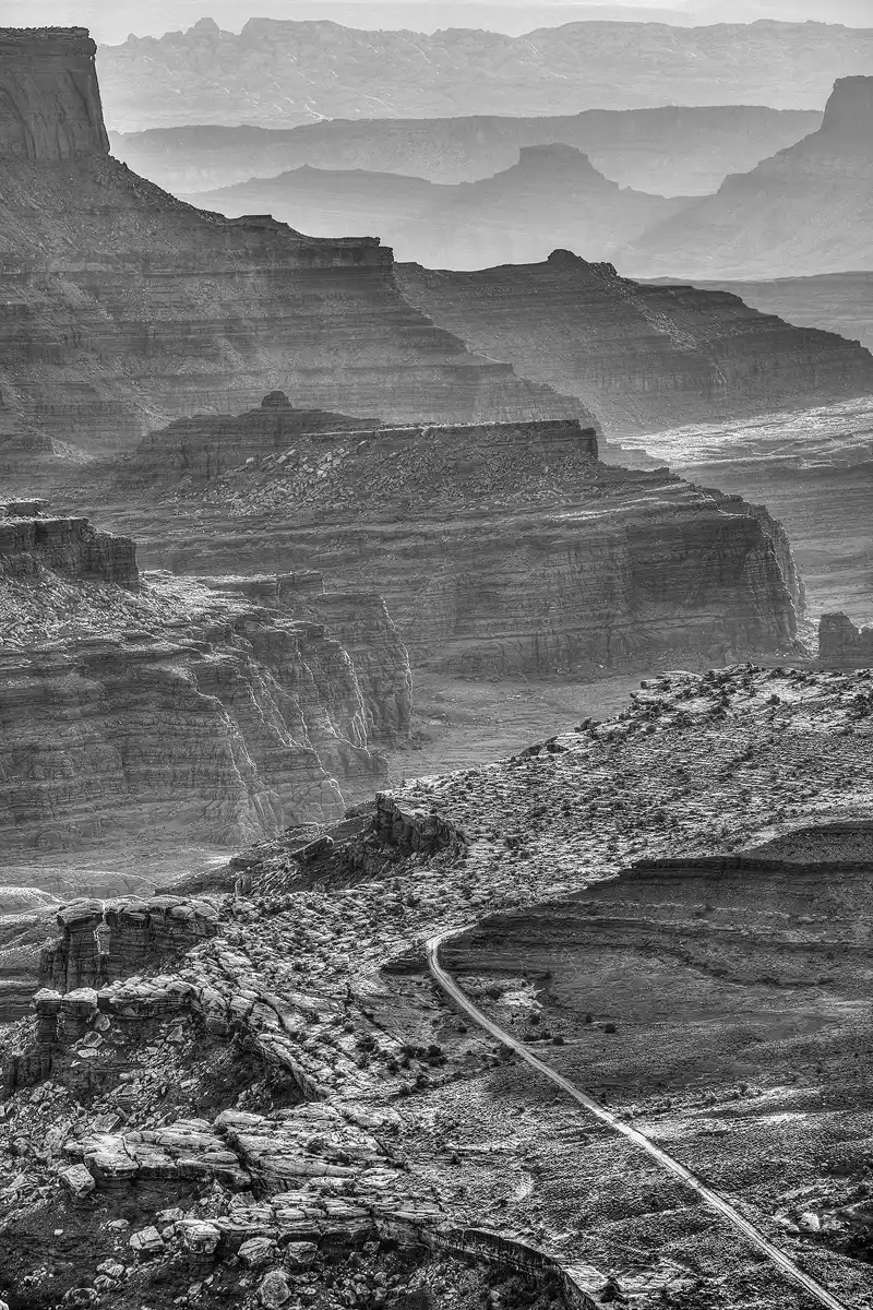 Vertical, black and white image taken with a long lens from a high mesa in Island in the Sky district of Canyonlands National Park. The upper two thirds of the image shows nine layers of mesas and ridges fading into the far background, higher in the image. The bottom of the image shows a section of a mesa top, it's rocky cliff edge on the left and a few undulating hills on the right. A dirt lane emerges in the lower right corner of the frame and angles up and to the middle where it disappears as it makes a right turn around a hill.