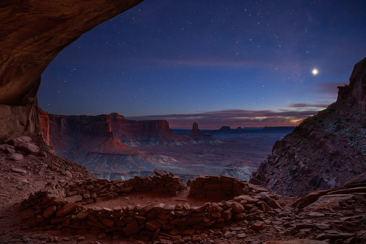The False Kiva is set in a shallow cave with a large arched opening on the side of a cliff face. This kiva is nothing more than a circular rock wall about two feet tall and fifteen feet in diameter positioned at the opening of the cave. This image was taken from the backside of the cave looking out over the kiva and showing the spectacular view of desert cliffs, buttes and mesas fading into the distance. What really adds to the appeal of this image is the time of day it was taken. It is well past sunset, but there is still blue in the mostly clear sky and many stars are shining in it.