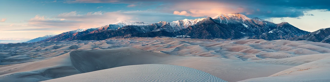 Wide panorama, taken at sunset, pointing north, from the tallest dune in Great Sand Dunes National Park. In the foreground are ripples in the sand of the closest dune and then layer after layer of sand dunes running into the distance and about half way up the frame. The top half of the image shows a long ribbon of the Sangre De Cristo mountains laden with winter snow and their cloud shrouded peaks receiving the last light of day.