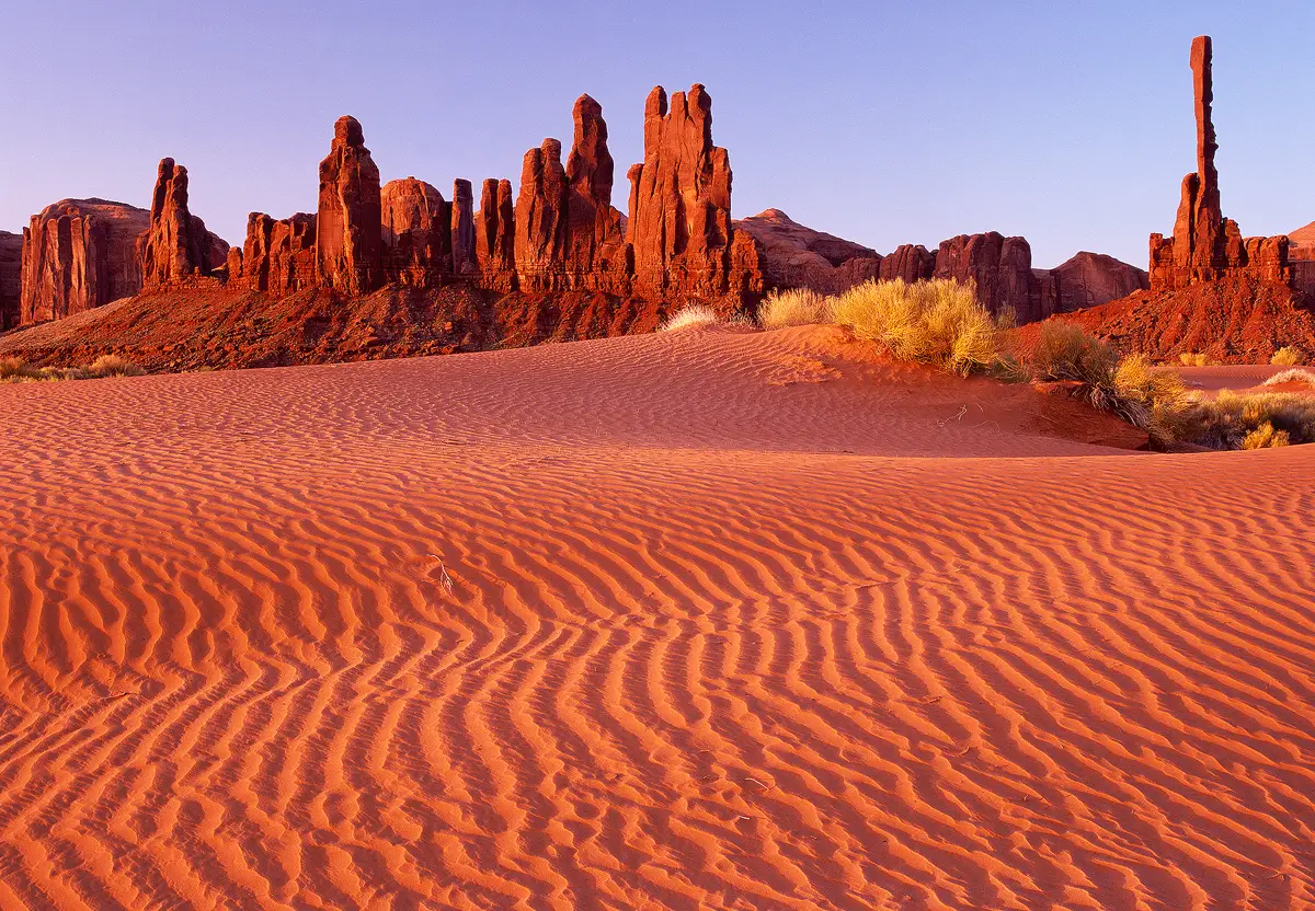 The Totem Pole is a lone rock spire in a series of rock spires in the desert landscape of Monument Valley, Arizona. The bottom two thirds of this predawn image shows sand ripples leading to the background, rock spires in the upper third of the image. A warm glow from the horizon, to the left, out of the scene, has caused the red sand and red rock spires to glow almost orangish.