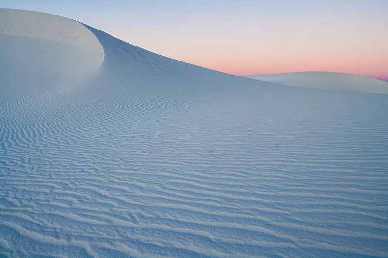 This stunning image of the White Sands National Park captures the ethereal beauty of New Mexico's dunes in the soft light of predawn. A large dune curves down from the upper left, filling the foreground with rippling sand. The interplay of light creates a striking contrast: the backside of the sand ripples, bathed in cooler blue tones, while the front sides, facing east, glow with warmer hues. In the background, another dune rises, leading up to a sky that transitions from a pinkish-purple at the horizon to a serene blue higher above. Captured by Andy Cook, this peaceful yet dynamic scene highlights the dramatic landscapes of White Sands at sunrise.