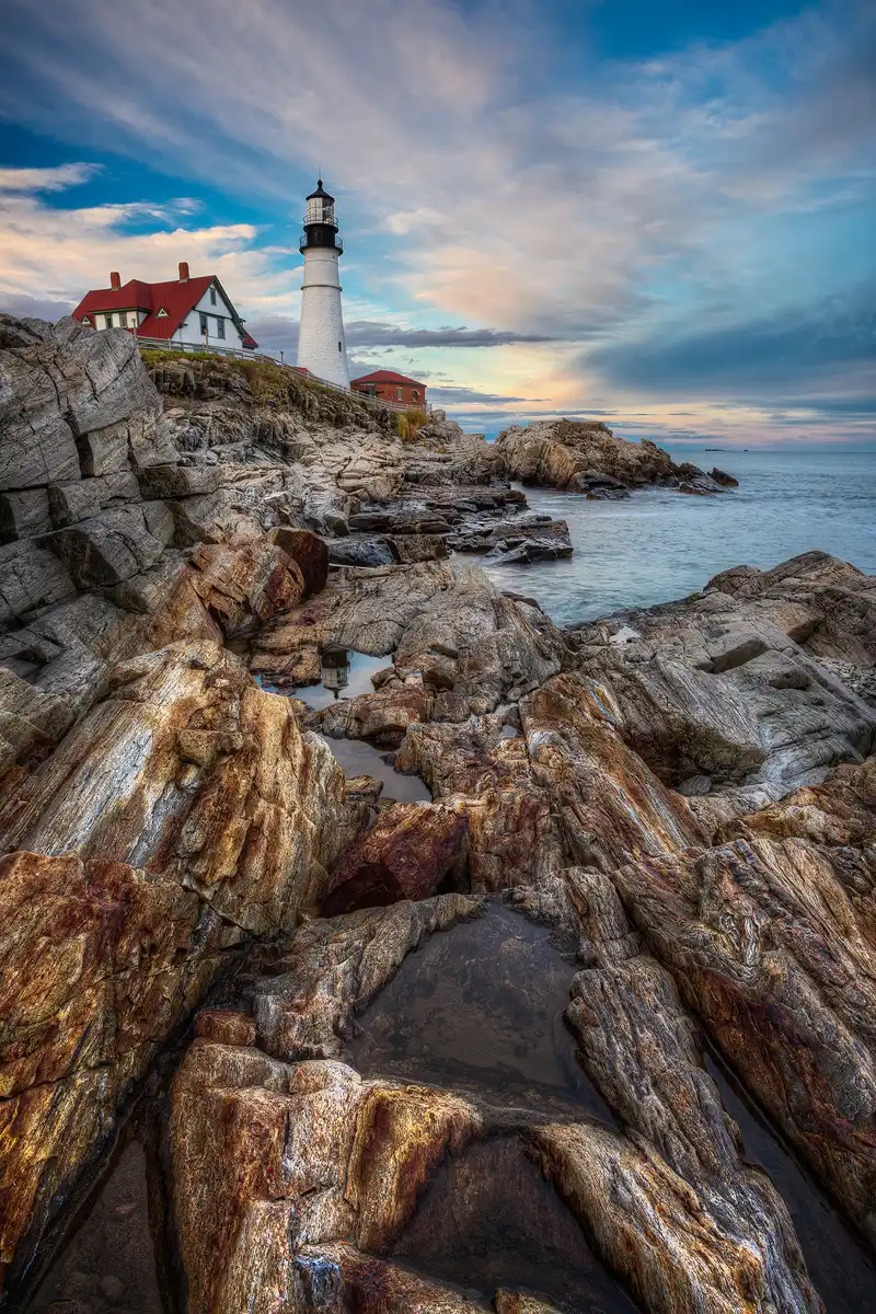 Portland Head Lighthouse at twilight with striated clouds and rocky coast.