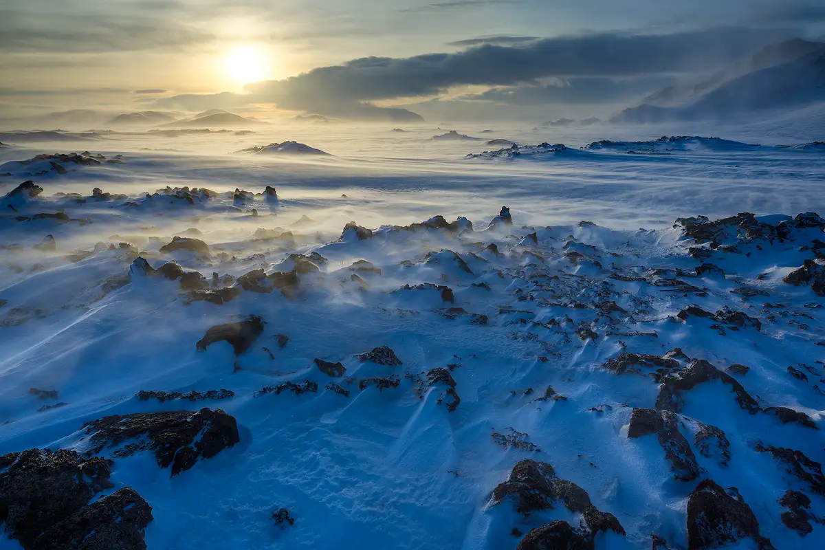 The bottom half of this winter image shows wind driven snow being pilled up toward the camera behind rocks and small piles of rocks, this area is still mostly shaded and has a blue cast. Higher in the frame, snow is being blown across an area with less rock piles then further back, small hills then larger ones arise and are partly obscured by blowing snow. The sun is low on the horizon and is positioned on the left side of the upper fifth of the image. It's behind so much haze and thin clouds that it's just a large glowing yellowish blob.