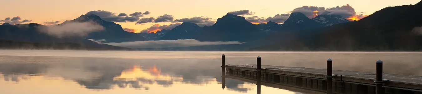 A light mist has formed over the calm mountain lake in the lower half of this panorama. A dock enters the bottom, right corner of the frame and marches out into the lake and ends about halfway into the frame partly obscuring the misty reflection of distant mountains. It's dawn and some of the puffy clouds clinging to the tops of the back lit mountains glow orange and show in the reflection.