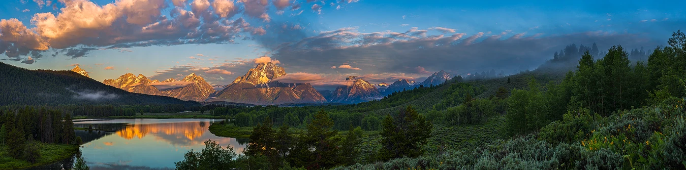 In this panorama only clouds and the tops of dramatic looking, background mountains are receiving warm, dawn light, everything else is still in shade. The high vantage point allows the viewer to look out and down to the calm water of Oxbow Bend on the Snake River in the bottom left of the frame. The water reflects the lit mountains behind. The low atmosphere is full of moisture as evidenced by clouds at many elevations including puffy clouds in the sky and a few mountain peaks beginning to have fluffy clouds condense around them. Also, partway down some mountains a thin band of clouds exists. The right side of the image shows a foreground of sagebrush and yellow flowers giving way to a mid-ground of stands of trees and sagebrush fields on an undulating hillside whose highest reaches are partly shrouded in fog.