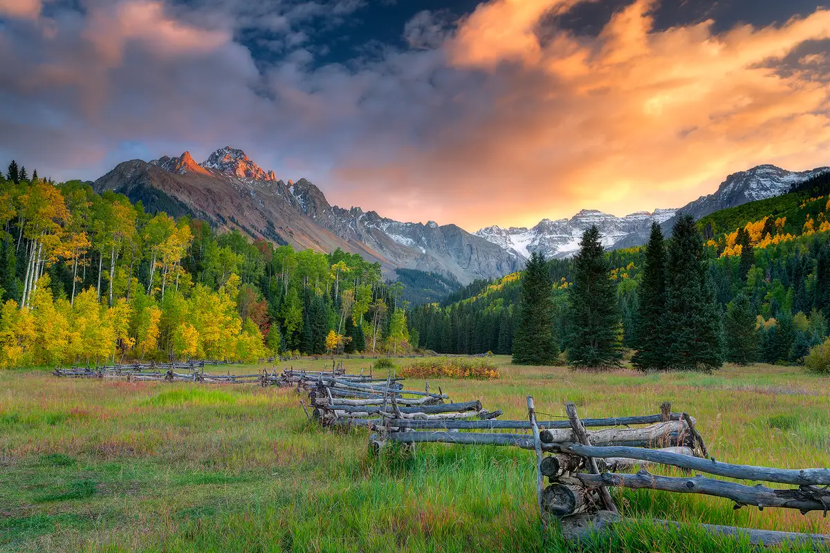 The bottom third of this image shows an old, zigzag fence entering in the bottom right corner of the frame then moving left across the frame and away into a meadow, eventually it curves right and becomes very small, disappearing in the middle of the frame. The middle third of the image reveals a beautiful autumn forest of colorful aspen and spruce trees and above it looms the rocky, craggy slopes of Mount Sneffels and it's wide cirque. The top of Mount Sneffels is glowing orange with the day's last sun rays and it is positioned in the upper left power point of the image. From there the cirque moves right, across and out of the frame. The upper third of the image is a partly cloudy sky ablaze with yellows and oranges.