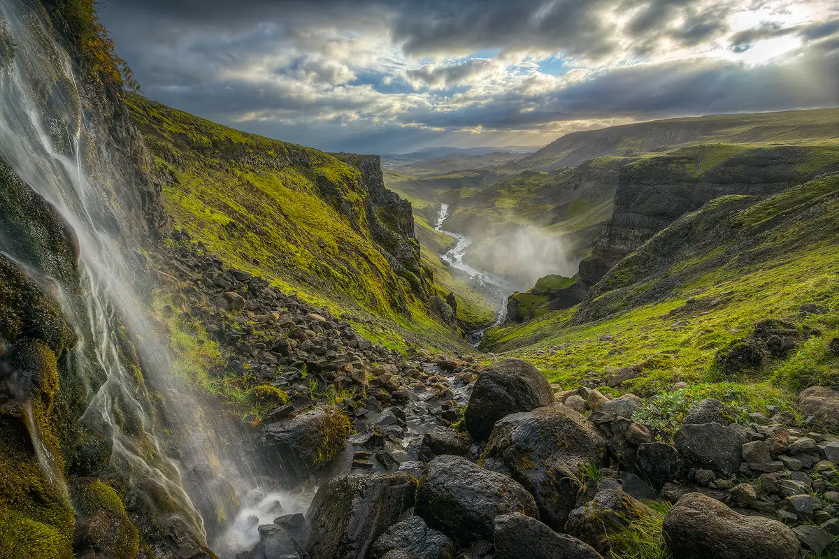 A small waterfall flows down from the upper left of the image and splashes on rocks in the foreground, on the bottom, about one third of the way in from the left. From there the stream flows down and away through rocks toward the middle of the frame before it disappears over a v shaped cliff. Just above from where it disappears, In the far distance, a river flows up and away from the middle of the frame and turns out of sight around a distant bend. Both sides of the close stream and distant river have green meadows of short grass, rocks and cliffs. The uppers part of the image shows the sun breaking through clouds clearing after a storm.