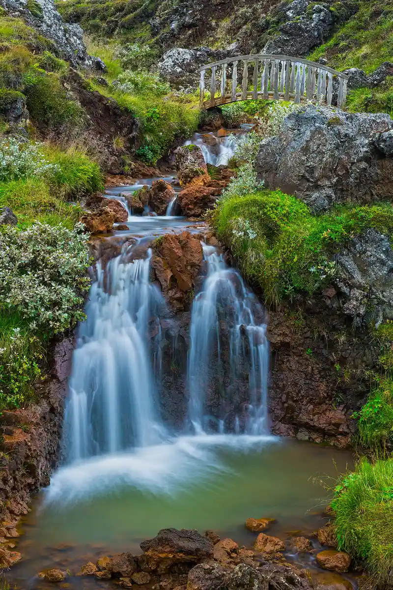 A small, foreground waterfall is in the bottom half of this image. It splashes into a greenish pool that is almost as wide as the frame. The rocks around the pool and the ones exposed by the stream are orangish. In the upper half of the image, the stream before the waterfall leads the eye up hill, away and to the right, to a small, arched wooden foot bridge. Both sides of the stream have green clumps of short vegetation and large rocks.