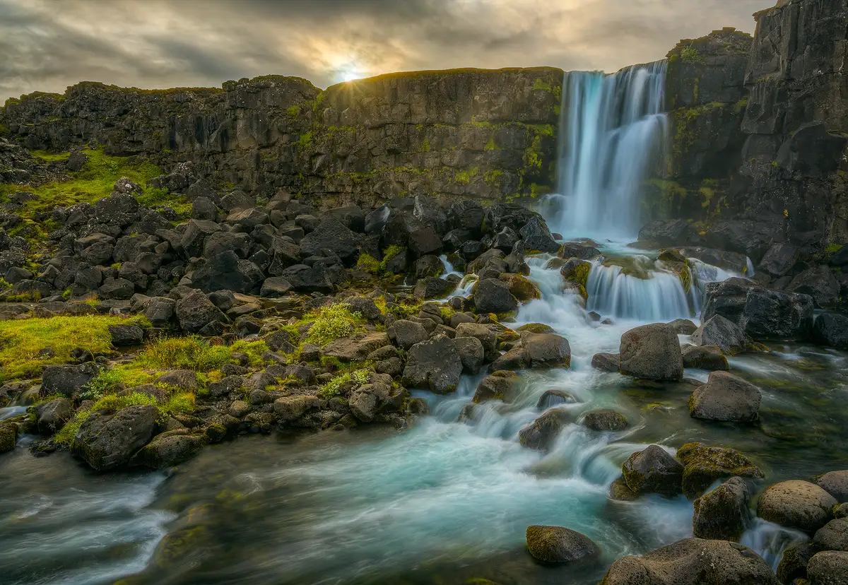A thirty foot cliff emerges from the upper right in the image and angles slightly down running all the way across and out the left side. Above it is a stormy sky and slightly left of the middle the sun is poking through just enough to cause a sun star at the top of the cliff. Blue-green water spills over the cliff in the upper right area of this image. The stream below continues to cascade around and over boulders and flows right in the frame before curving to the left and exiting in the bottom left corner. The far bank of the stream is littered with boulders and areas of short, green vegetation that lead back the cliff.