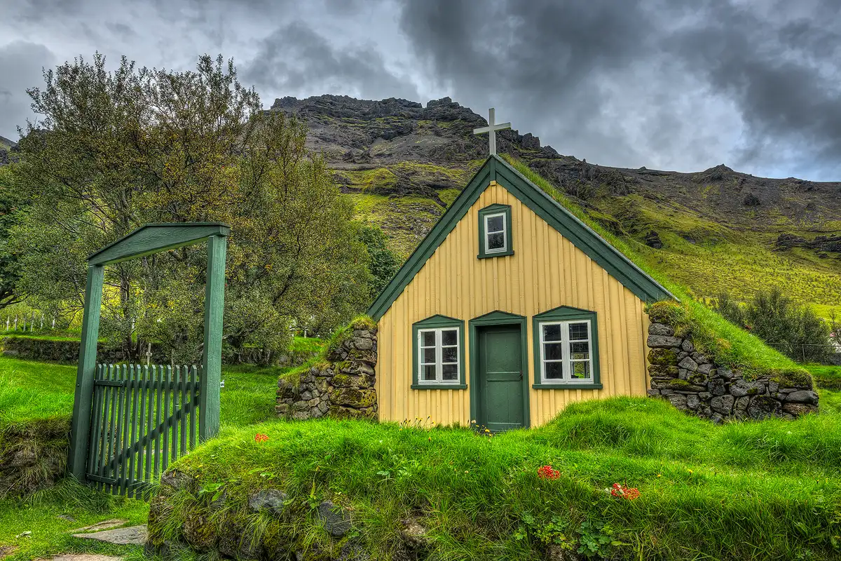 Hofskirkja turf church is in Southeast, Iceland. The church and it's rural setting have a fairy-tale like quality. A layer of grass spills over the top of the small building. The building's front is board and batten and painted a subdued yellow. The front door and trim are painted forest green. A few steps in front of the church and lower in the frame, is a rock wall that is totally over grown with green moss. Along the top of the wall there is thick, green grass with a few clumps of red berries placed on it. To the left of the church a path leads to a break in the rock wall where there's a forest green, wood fence gate. The dark side of the fairytale seems apparent in this image. In the background a stormy sky and darker mountain loom over the scene with ominous overtones.