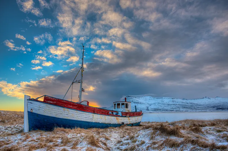 In this serene scene, an old wooden fishing boat rests near the shore of an eastern Icelandic fjord, surrounded by long brown grasses lightly dusted with winter snow. The boat, once brightly painted with three horizontal stripes—red on top, white in the middle, and blue at the bottom—now shows the weathered charm of time and use. Across the fjord, majestic snowy mountains rise toward the morning sky, which is filled with beautiful clouds catching the first light of day. This peaceful landscape, captured by Andy Cook, showcases the rugged beauty of Iceland and the timeless presence of the fishing vessel against a dramatic natural backdrop.