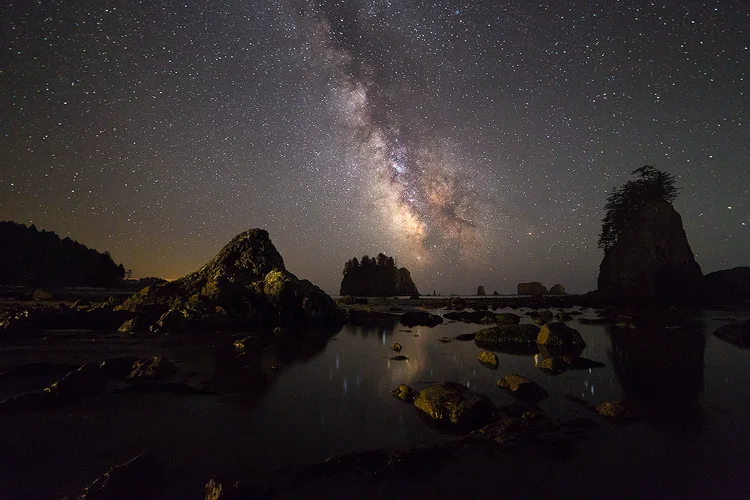 Night photo of Second Beach in Olympic National Park. The Milky Way rises up, at a left angle, into the clear night sky. The bright, galactic core hovers just above the horizon. In the mid-ground, left and right of the core, tree capped islands jut into the night sky. In the foreground, reflecting the Milky Way, is a large pool of calm water with a scattering of rocks.