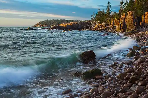 A wave splashes over small, round boulders on the curving shoreline as morning light warms the cliffs above Thunder Hole Beach. This kind of image may be photographed during our Acadia National Park photography workshop.