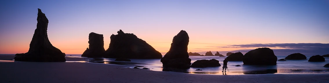 Sunset silhouette of a photographer and sea stacks on an Oregon beach, reflecting in the surf. Landscape photography workshop scene.