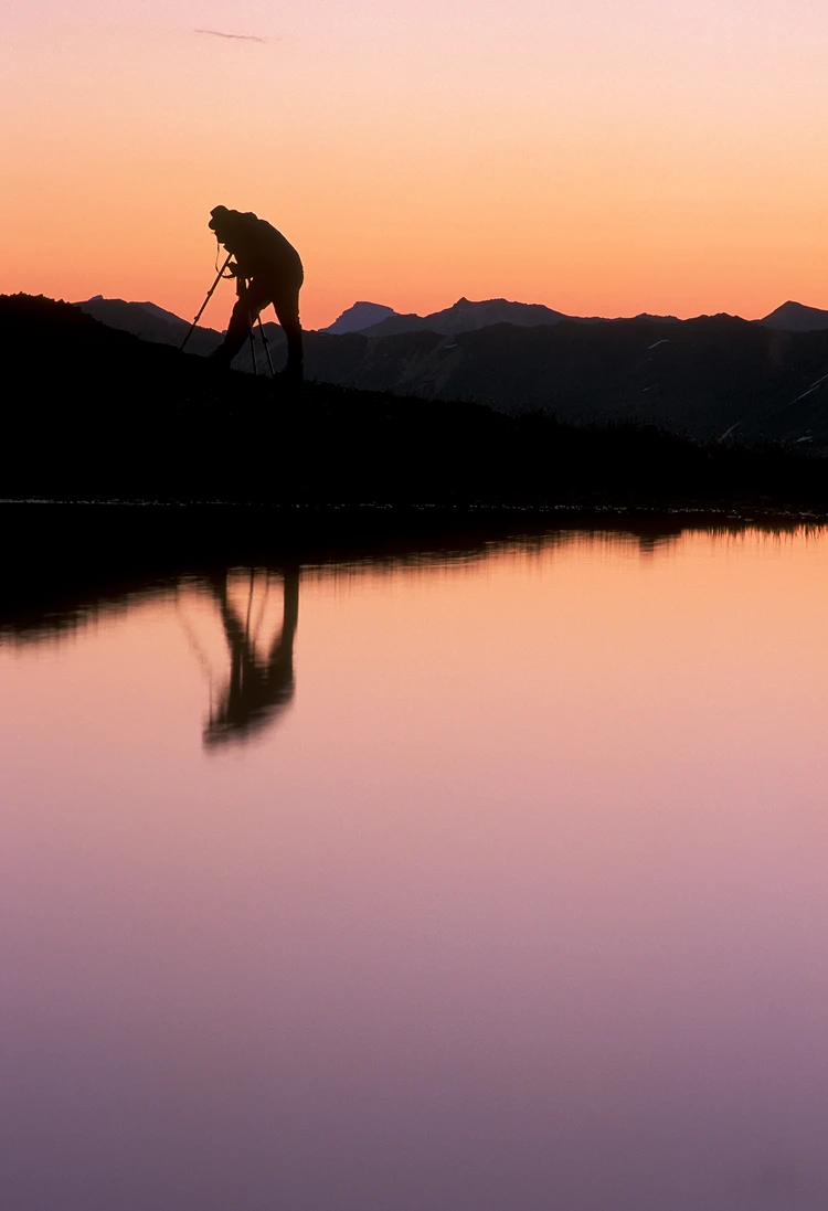 Photographer silhouetted at dawn, capturing a landscape reflection.