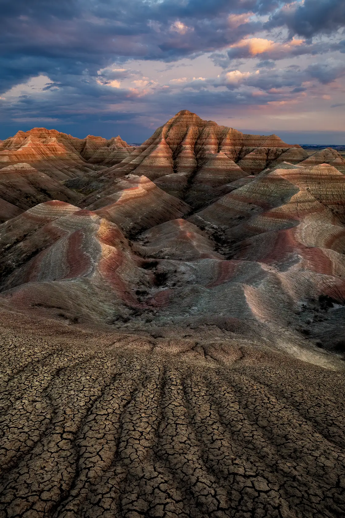 Cracked mud erosion patterns leading toward colorful Badlands formations under dramatic evening storm clouds in Badlands National Park.