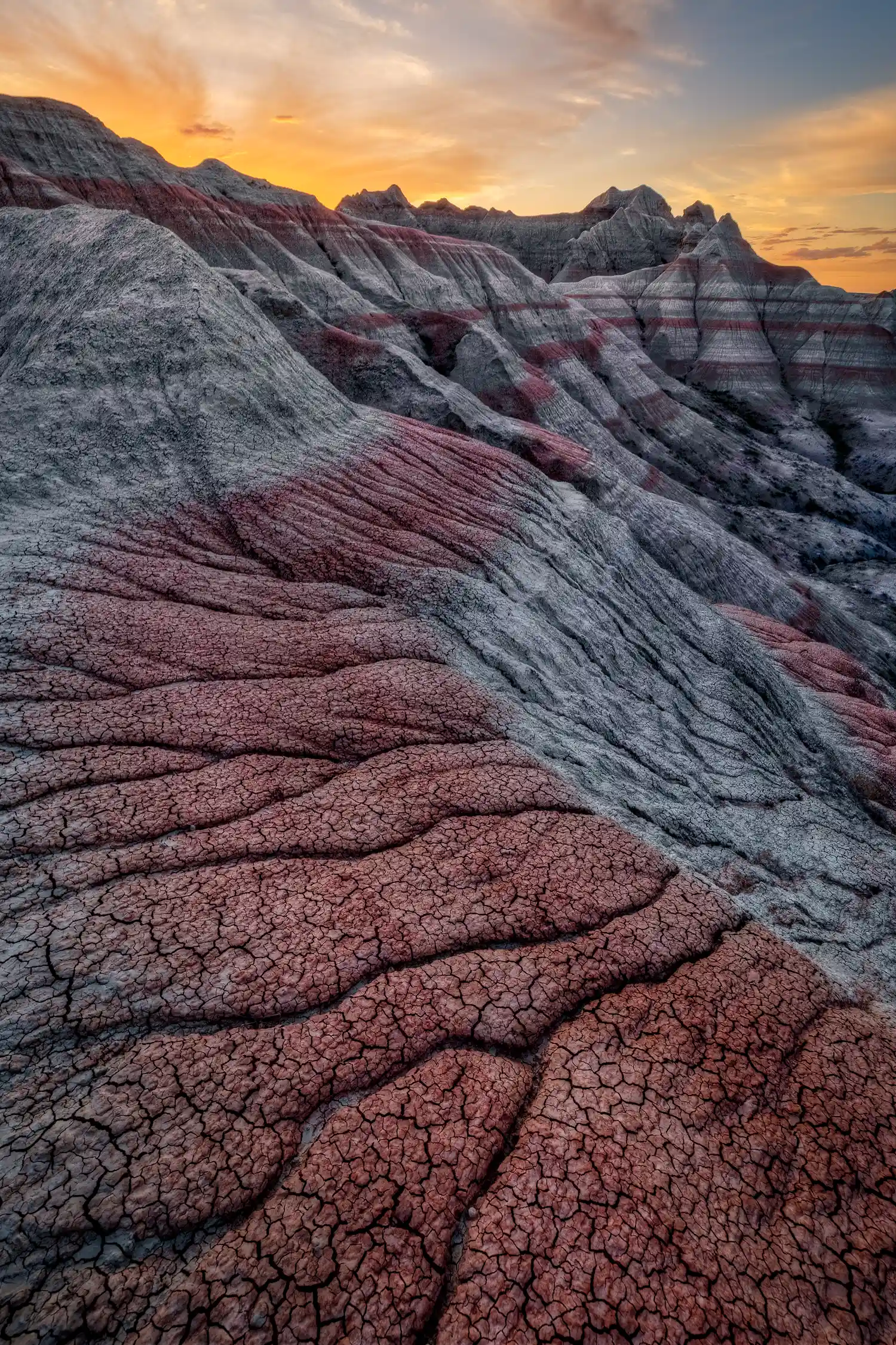 Red soil stripe leading to layered rock formations under a sunset sky with orange clouds in Badlands National Park.