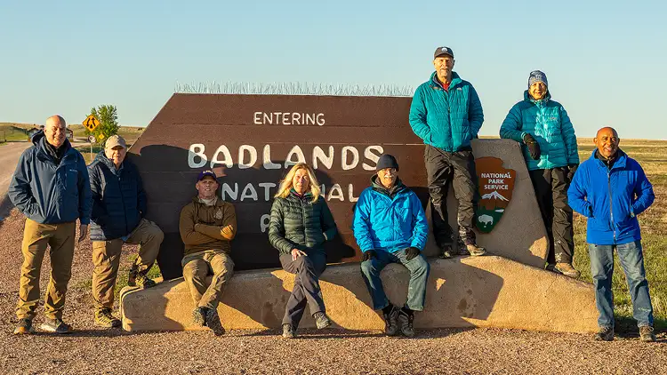 Photography workshop group at Badlands National Park sign, looking tired but fulfilled after days of shooting, some seated, some standing, in soft morning light.