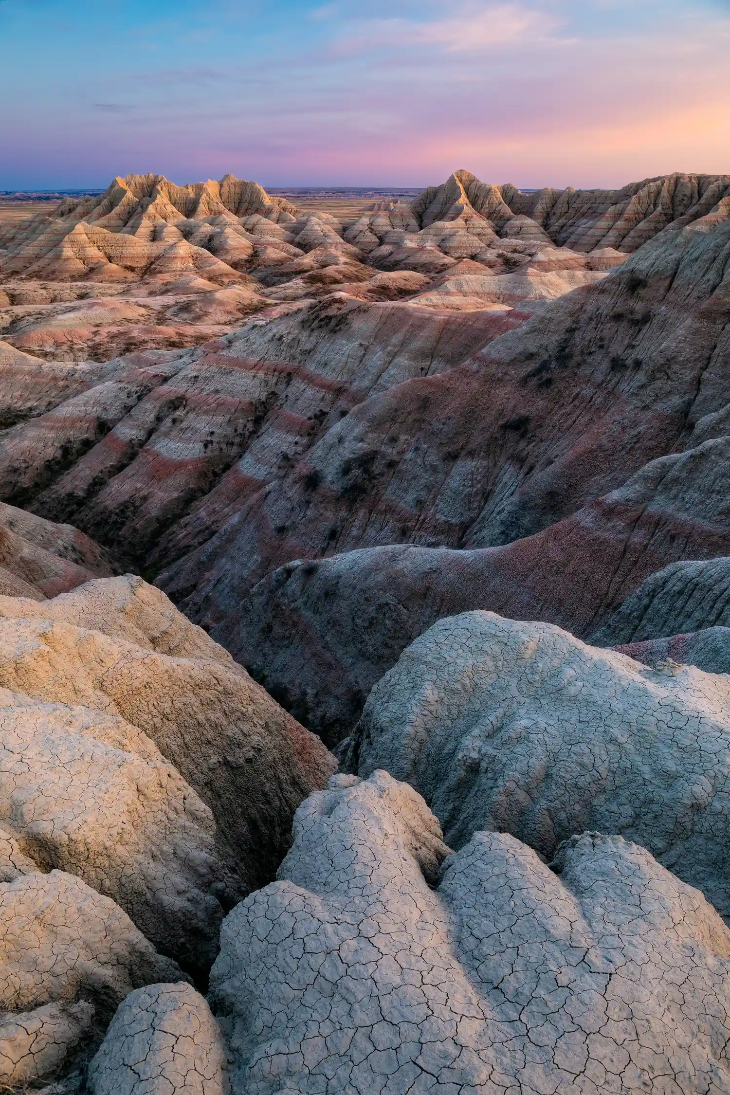 Mud cracks leading to a ravine and distant mountains with red and white layers in Badlands National Park at sunset.
