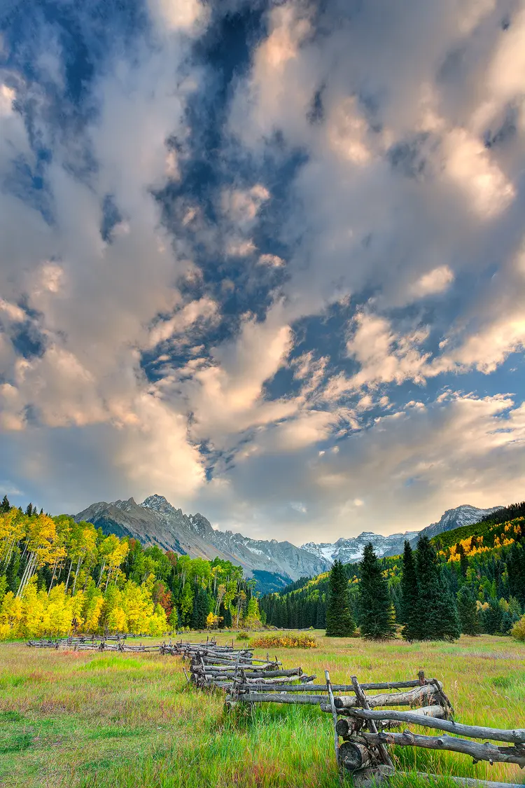 A weathered zigzag fence leading towards a vibrant aspen grove and the majestic Mount Sneffels under a dramatic, cloud-filled autumn sky in Colorado.