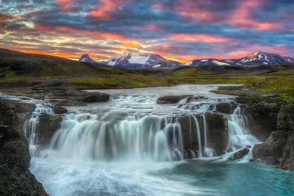 Warm, orange and yellow sunrise colors dance across the partly cloudy sky. A river flows away from distant, snow capped mountains and cascades over a rock shelf in the foreground creating a wide waterfall flowing out the bottom right of the frame. Iceland is a land of simple beauty and powerful contrast and is a treasured destination for landscape photographers.