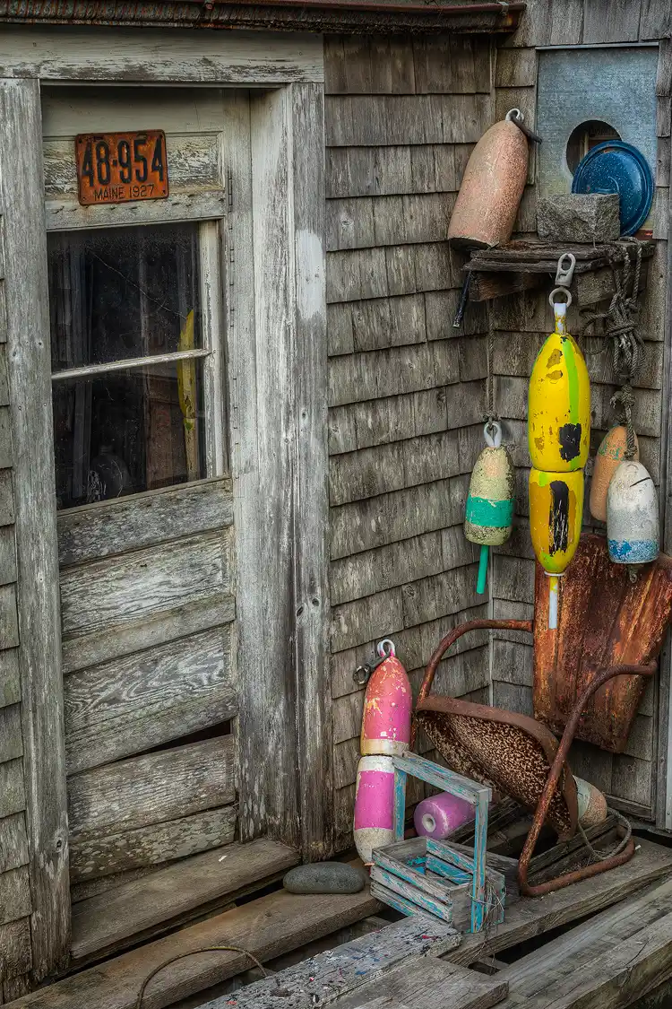 Photography location; Vertical Maine coast image of a severely weathered door on an old lobster shack. Lobster buoys and a rusty chair are aside the door.