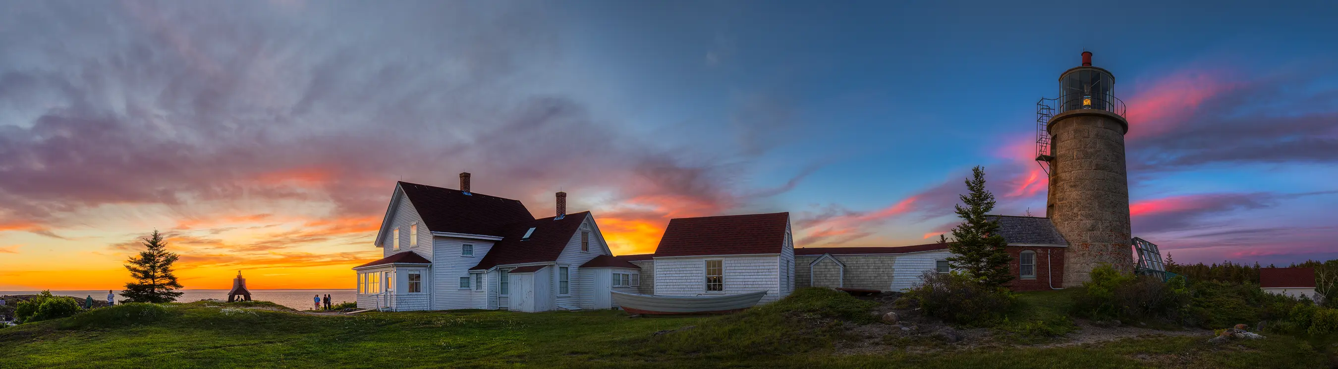 Panoramic sunset view of Monhegan Lighthouse with a vibrant sky on the Maine coast.