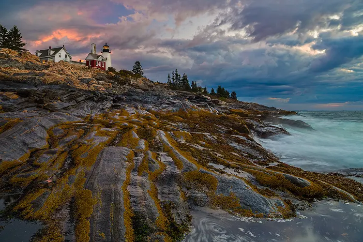 Workshop location; The Atlantic ocean swirls around shore rocks on the far right side of this image. In the bottom powerful leading lines are created by barnacles preferring a certain zone in the striated granite. The lines lead up and to the left, to the Pemaquid Lighthouse. A red and white set of buildings with the light still shining in the early morning. Above it, stormy clouds partially glow orange with dawn's first light.