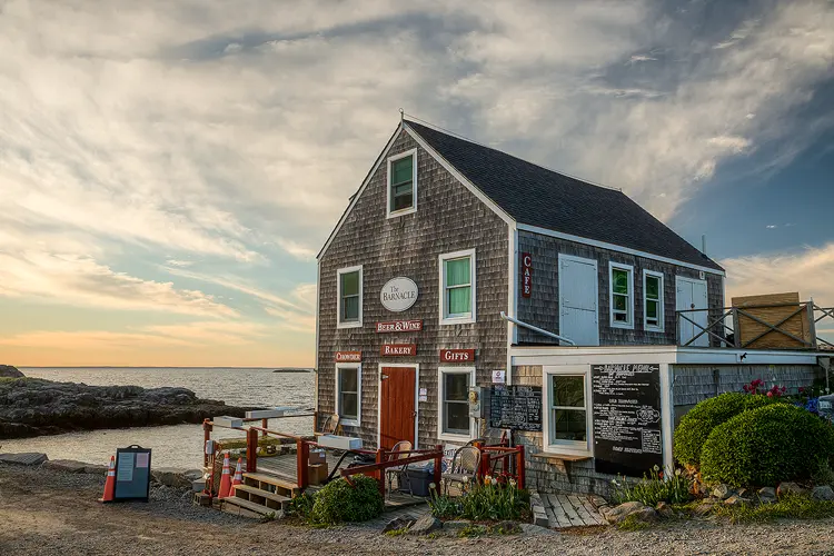Photography location; This cafe and gift shop at waters edge on Monhegan Island is a classic Maine coast building. It is two stories tall, with weathered, gray shake siding, white trim, dark roof and a red door. It looks fantastic in late day with muted light and clouds streaking above.