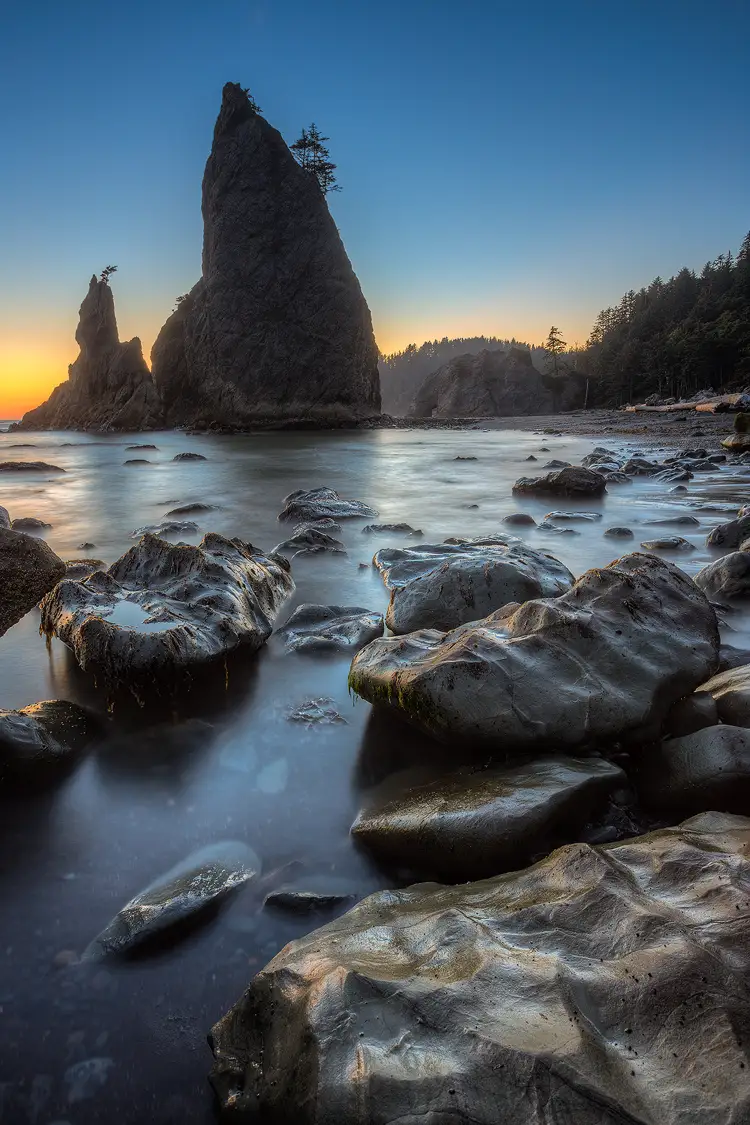 Photography location; The sun has set, but the sky still glows orange and yellow near the horizon, transitioning to blue a bit higher. This vertical image shows the shoreline at Rialto Beach in Olympic National Park. Here tall rock spires, decorated by the occasional tree, point into the sky. In the foreground are large rocks partly in the surf which is made smooth by a long exposure. The sculpted rocks form a visual a path back to the spires.