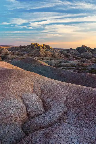 Eroded hill with dendritic and mud crack patterns in the foreground, dramatic Badlands mountains with red and white layers in the background, illuminated by sunset light. Photo by Bob Hart.