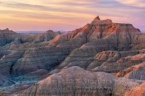 Rugged Badlands mountains with red and white layers under a colorful sunrise sky with warm sunlight. Photo by Bob Hart.