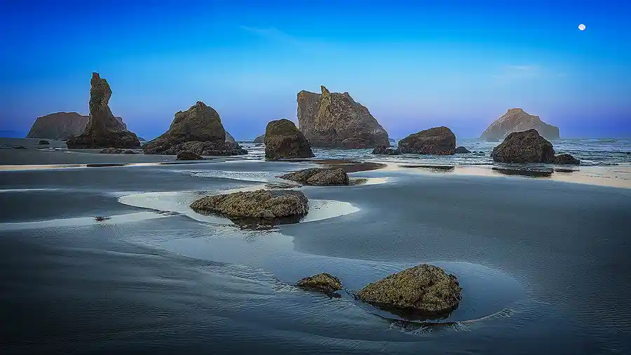 Panoramic sunrise photo of Bandon Beach with tide pools reflecting blue and pink hues, distinct sea stacks in the distance, and the moon in a clear sky.