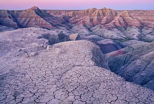 Badlands National Park at dusk, with purple-pink light illuminating eroded hills, mud cracks in the foreground, and red and white rock layers in the background. Photo by Cindy Beckett.