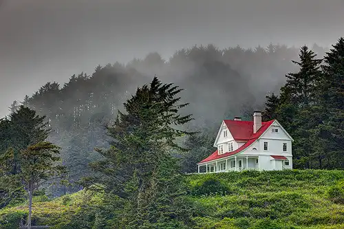 Heceta Head Lighthouse Keepers House on a misty, stormy day along the Oregon Coast, with green foliage and fog partially obscuring distant pine trees. Photo by George Casey.