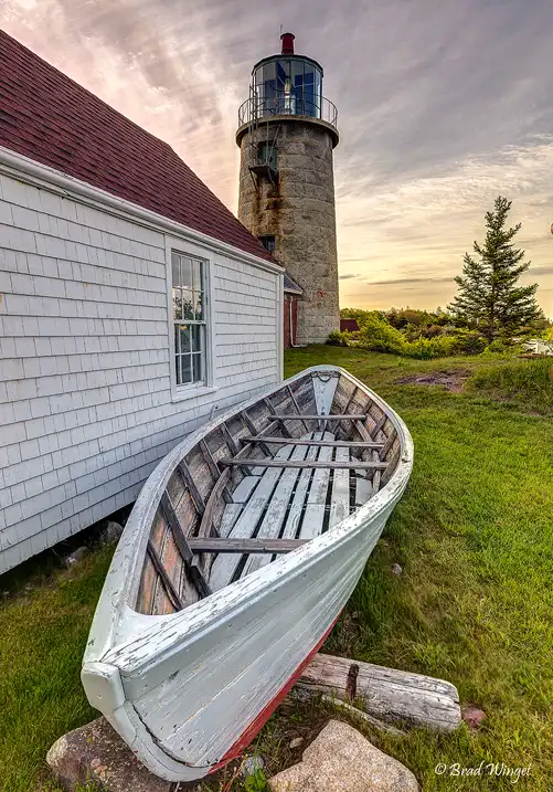 Weathered rowboat resting beside Monhegan Island Light beneath a colorful sunrise sky on Monhegan Island, Maine.