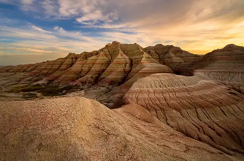 Badlands National Park at sunset, with colorful clouds in the sky, intricate mud cracks and eroded channels in the foreground, and red and white striped sediment layered hills in the background. Photo by Cindy Beckett.