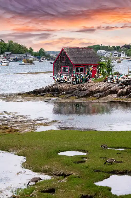Quiet tidal marsh leading to a weathered fishing shack trimmed in red beside a peaceful Maine harbor at sunset.
