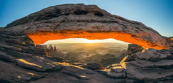 Sunrise panorama of Mesa Arch in Canyonlands National Park, with golden light illuminating the arch and rocky landscape. Photo by George Casey.