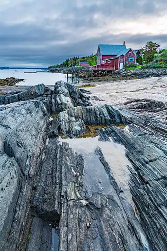 Red building and pier on Maine coast. Striated rock foreground. Morning light. Photo by James Gheida.