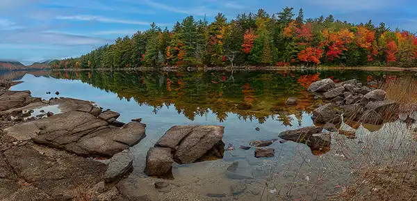 Predawn panorama of calm Eagle Lake in Acadia National Park, with granite rocks in the foreground and vibrant autumn trees reflected in the water. Striated clouds fill the sky, adding depth to the tranquil scene. Photo by George Casey.