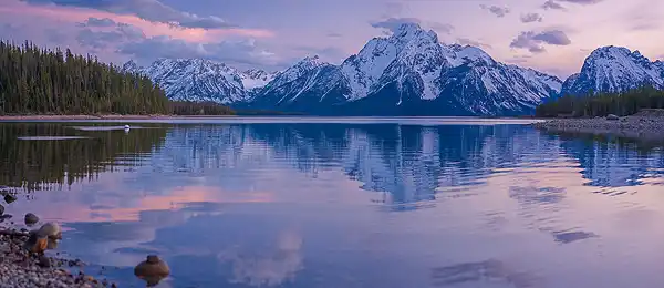 Post-sunset panorama of Jackson Lake, with Mount Moran's snow-capped peaks reflected in the calm water. Pink clouds add a warm glow to the evening sky. Photo by George Casey.
