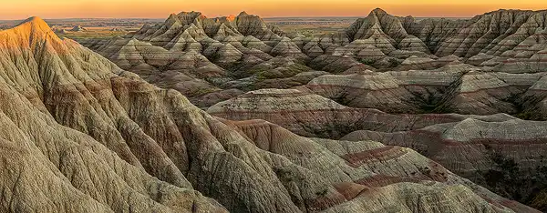 Panoramic view of sunset lighting the tops of eroded hills at Badlands National Park, with rugged mountains in the distance and red and white sediment layers. Photo by George Casey.
