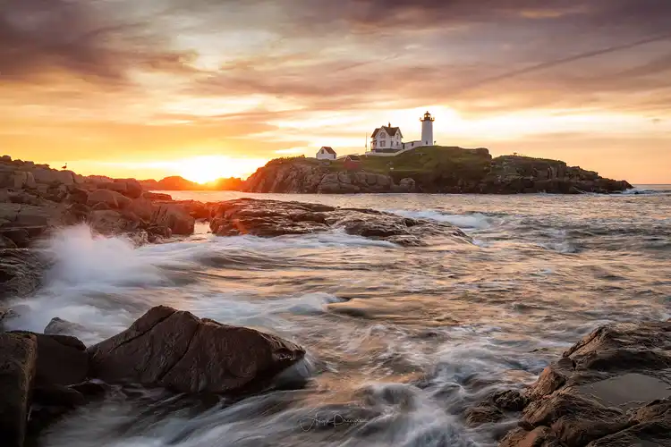 Morning surf rushing through rocky shoreline beside Nubble Lighthouse at sunrise on the Maine coast.