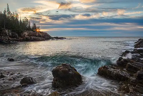 Sunrise at Acadia National Park, with a long exposure of curling waves, boulders on the beach, and golden light breaking through trees. Photo by George Casey.