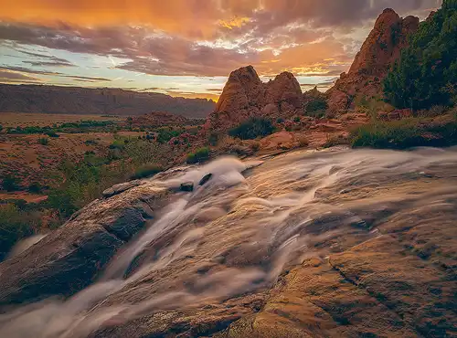 Sunset at Faux Falls near Arches National Park, with vibrant clouds, flowing stream, and rugged hills in the foreground. Photo by Cindy Beckett.