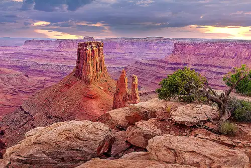 Sunset at Marlboro Point near Arches National Park, with vibrant clouds, large boulders, a juniper tree, and a distant mesa. Photo by Cindy Beckett.